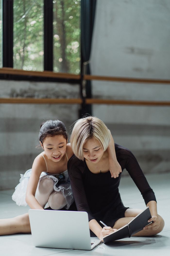 Smiling Asian ballerina sitting near coach taking notes and using laptop in dance hall
